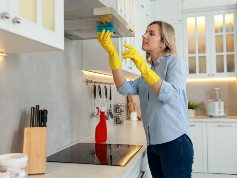 A woman wearing yellow gloves cleans a kitchen range hood with a sponge, showcasing her kitchen cleaning routine, with cleaning supplies visible on the counter.
