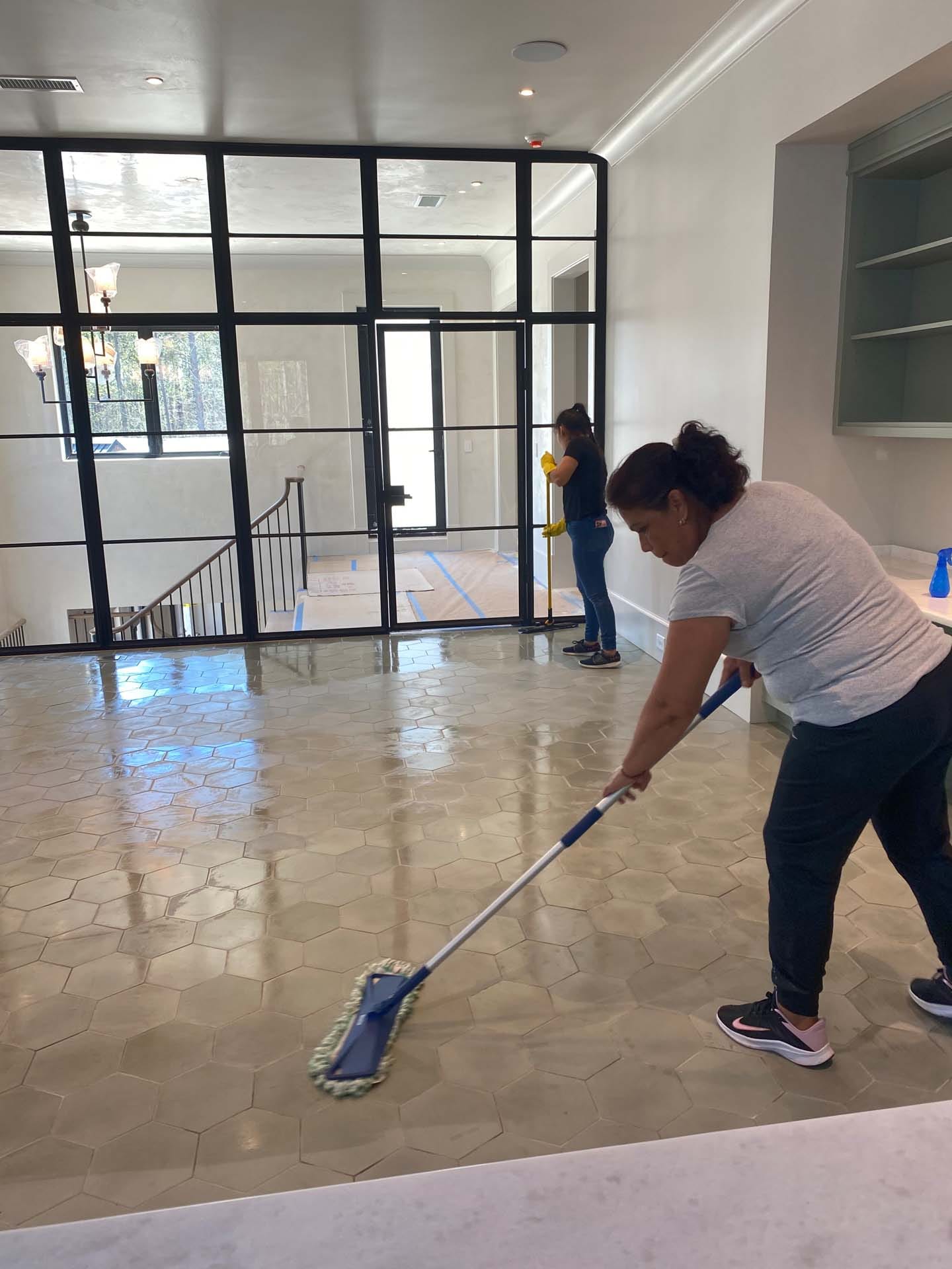 Two women performing post-construction cleaning, with one mopping a tiled floor while the other wipes glass doors in a spacious, modern room.