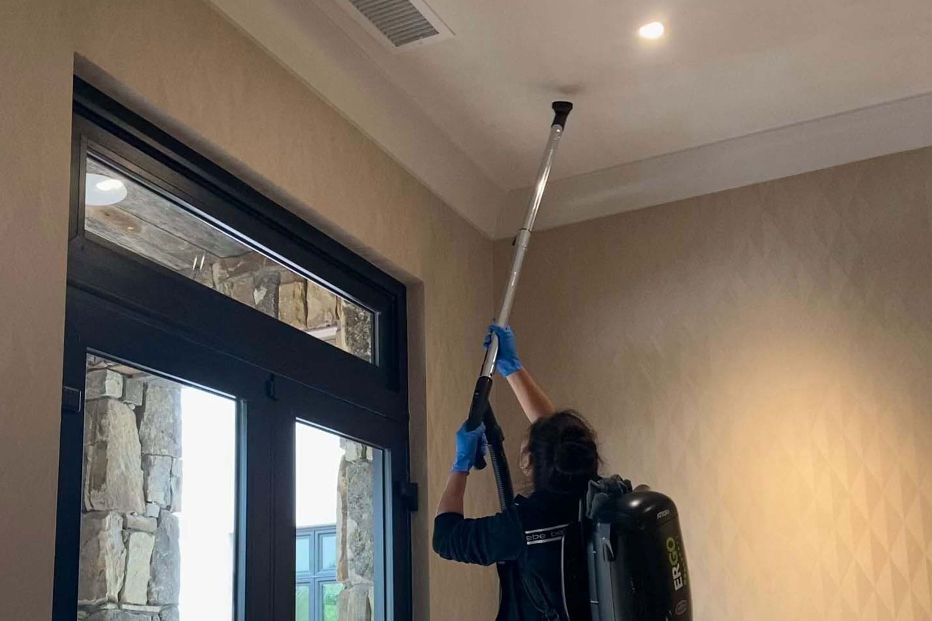 Person wearing gloves and a backpack vacuum performs post-construction cleaning near a ceiling corner using an extension pole in a room with beige walls and a large window.