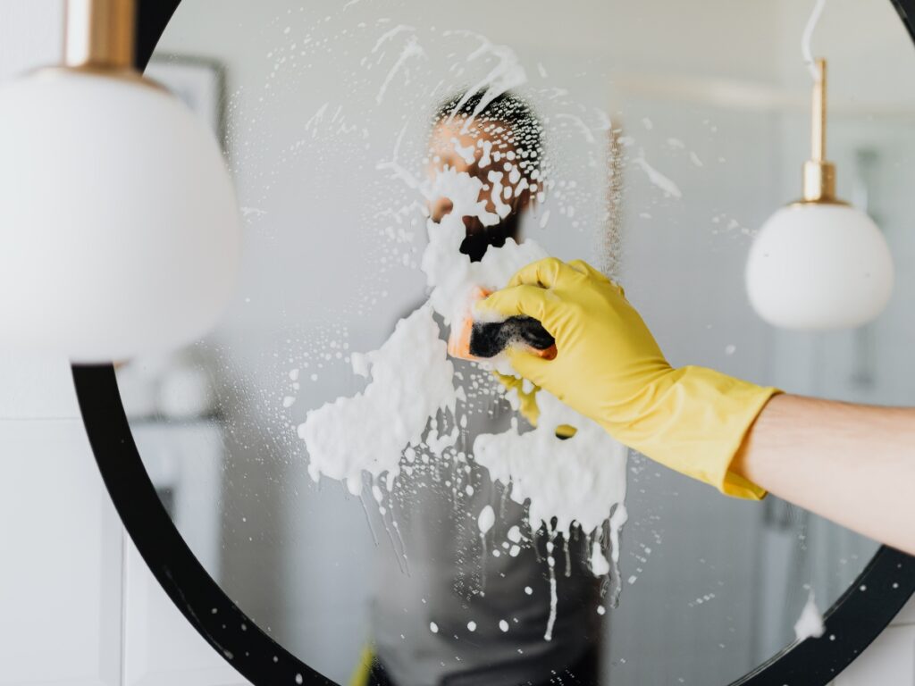 A person wearing a yellow glove uses advanced techniques to clean a round mirror with soap and a sponge; their reflection is visible in the mirror.