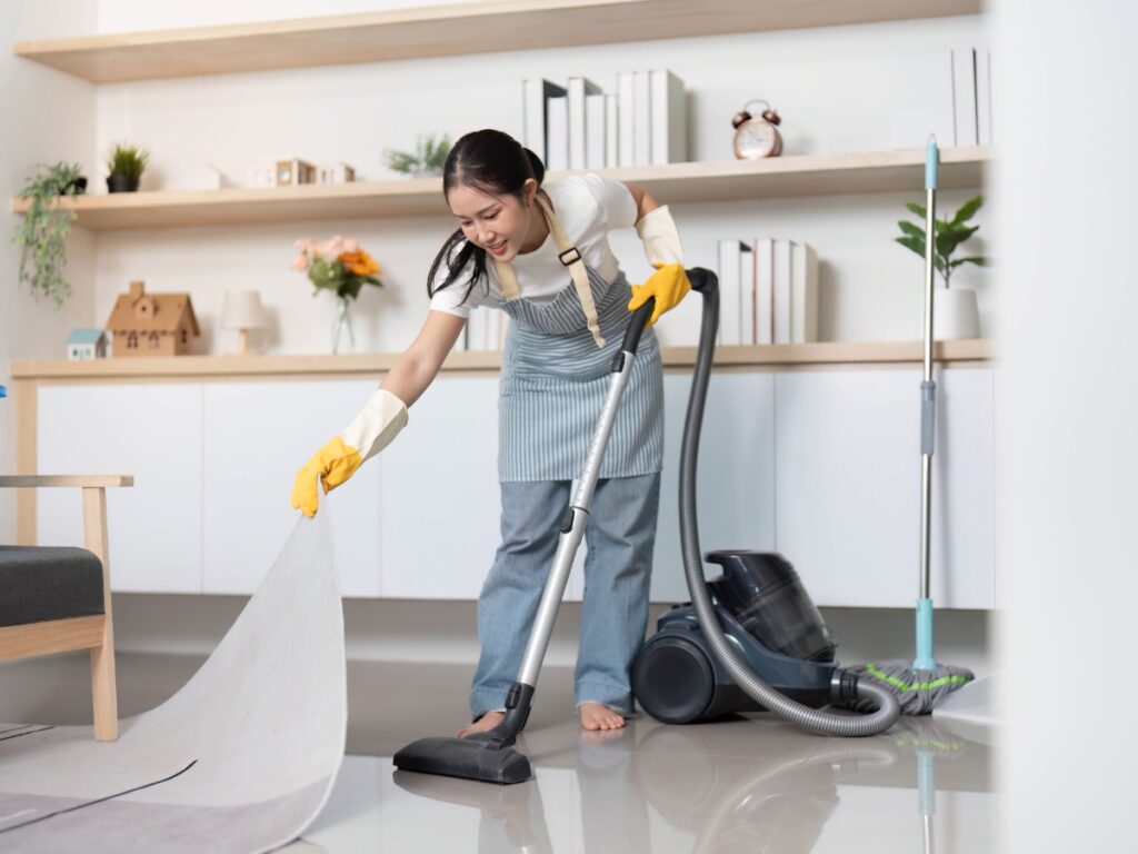 A woman using advanced techniques, wearing gloves and an apron, vacuums the floor while lifting a rug in a modern, tidy living room.