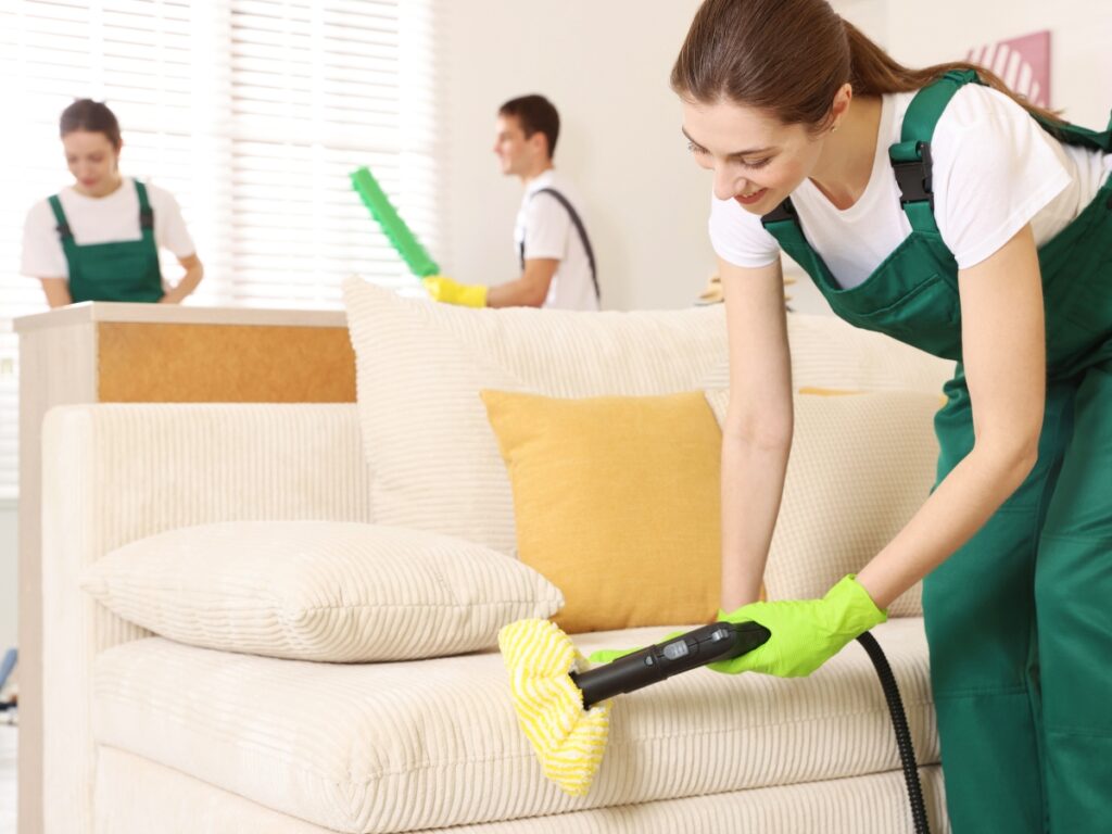 Three people in green uniforms clean a living room using advanced techniques; one woman uses a steam cleaner on a beige sofa while others dust and tidy in the background.